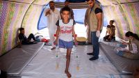 Abdel Rahman stands with crutches in his tent, while HI physiotherapist Mohammed looks on.